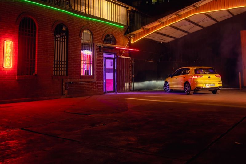 Rear view of a yellow VW Golf at night in a car park, the LED headlights illuminating a building.