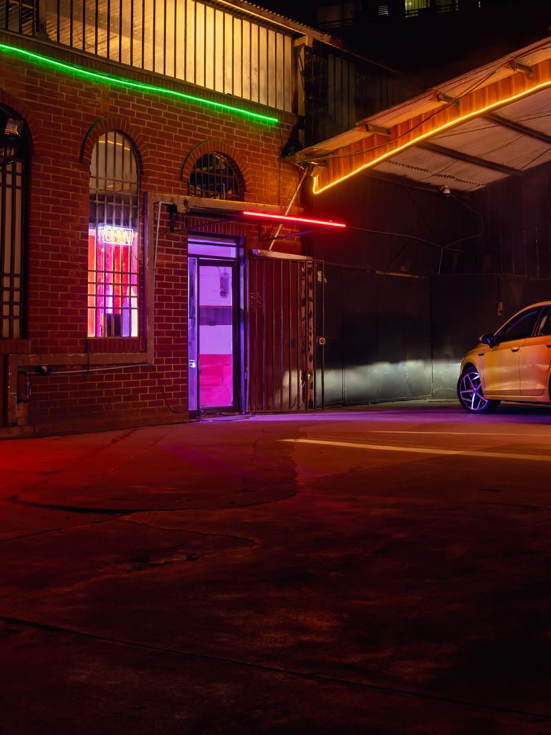Achteraanzicht van een gele Volkswagen Golf bij nacht op een parkeerplaats, waarbij de LED-koplampen een gebouw verlichten.