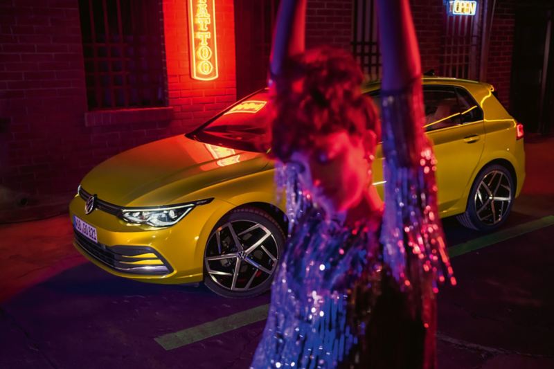 Gelber VW Golf steht nachts vor einem Leuchtschild, im Vordergrund eine glückliche Frau.