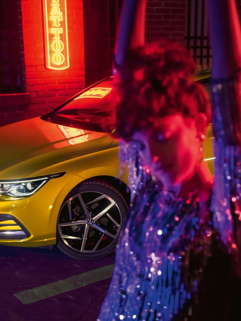 Gelber VW Golf steht nachts vor einem Leuchtschild, im Vordergrund eine glückliche Frau.