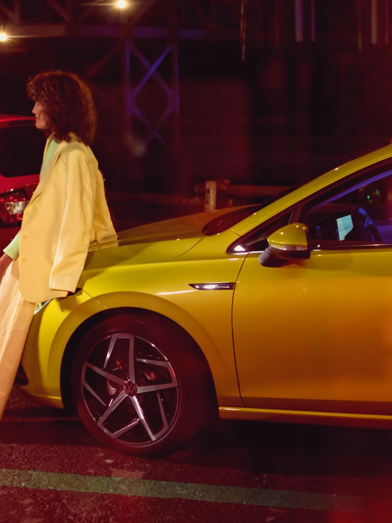 A couple leans chatting on the bonnet of a yellow VW Golf at night.