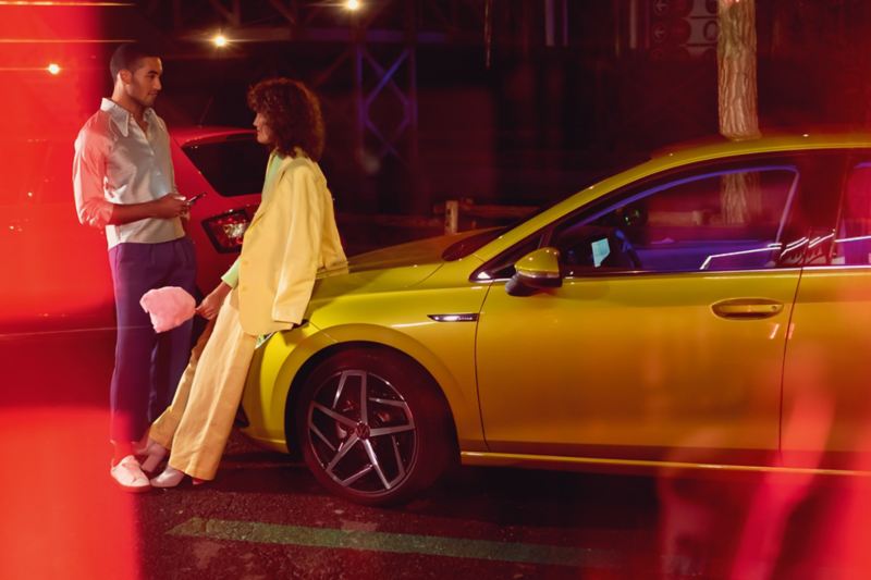 A couple leans chatting on the bonnet of a yellow VW Golf at night.