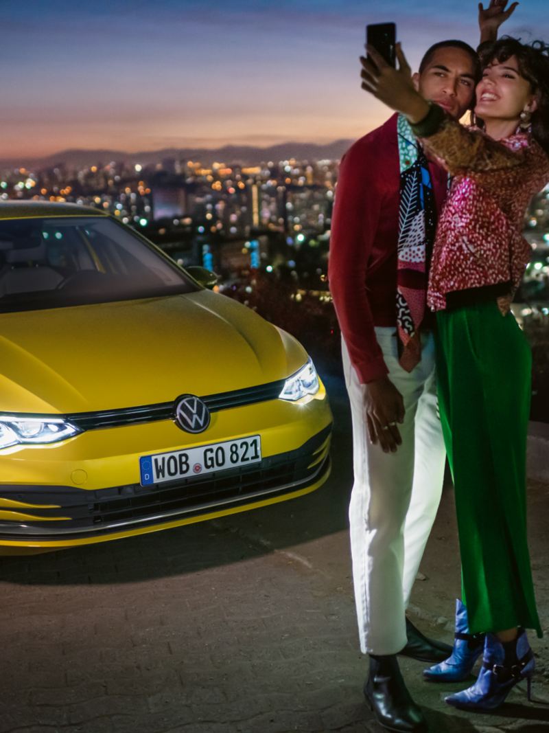 A couple are taking a selfie in front of a yellow VW Golf, in the background a valley with a city.