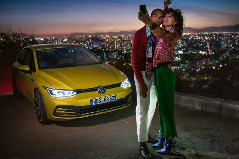 Un couple prend un selfie devant une VW Golf jaune, avec en arrière-plan une vallée et une ville.