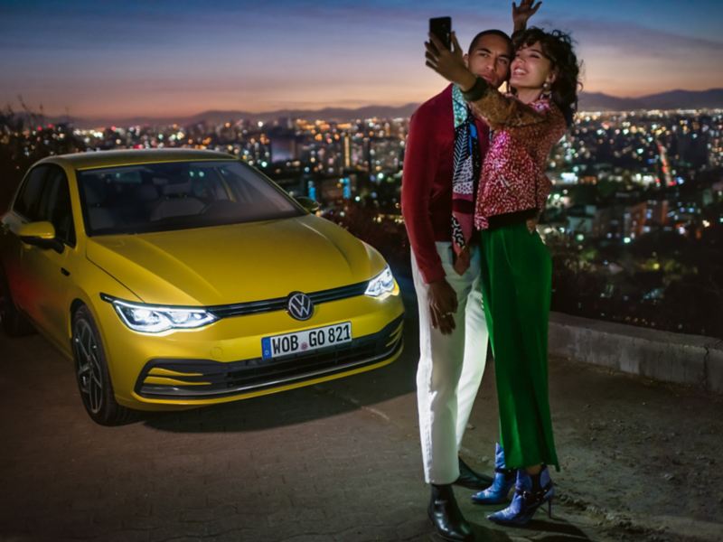 Ein Pärchen macht ein Selfie vor einem gelben VW Golf, im Hintergrund ein Tal mit einer Großstadt.
