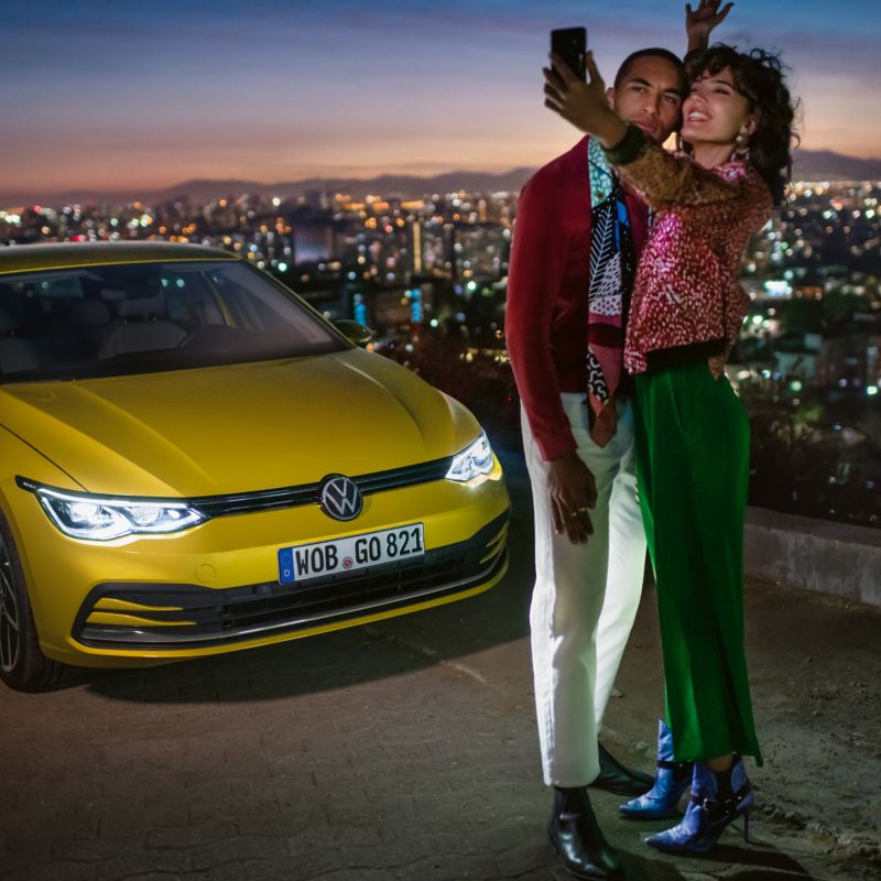 Ein Pärchen macht ein Selfie vor einem gelben VW Golf, im Hintergrund ein Tal mit einer Großstadt.
