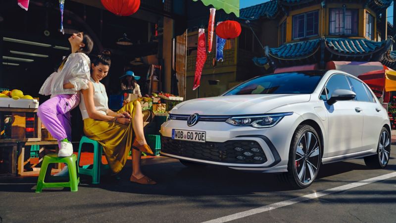 VW Golf GTE in a city centre with Asian features next to a vegetable stall where two young women are sitting.
