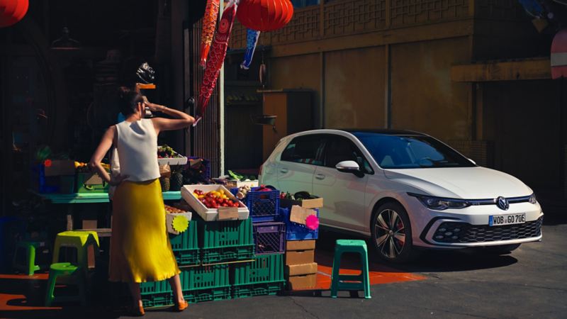 VW Golf GTE in bianco, vista laterale dal davanti, in piedi davanti a uno stand di verdure, di fronte c'è una donna