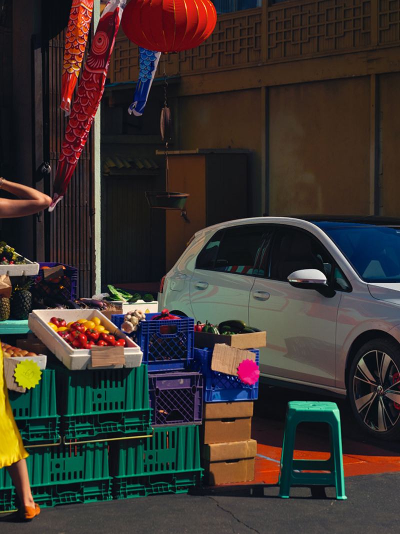 VW Golf GTE in white, front side view, stands at a vegetable stand, in front of it stands a woman
