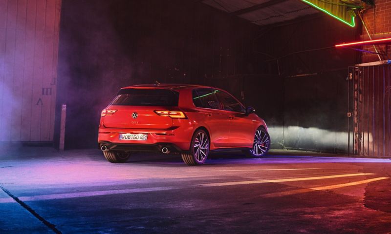 La VW Golf GTI rouge est garée de nuit sur un parking, les phares éclairent un mur, léger brouillard au sol.