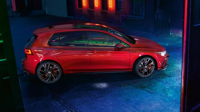 VW Golf GTI in red, side view, stands on a concrete square in front of a building with glowing signs at night.