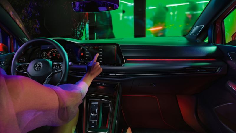 "Cockpit of the VW Golf GTI with red background lighting above the glove box and in the footwell, a woman using a touchscreen. "