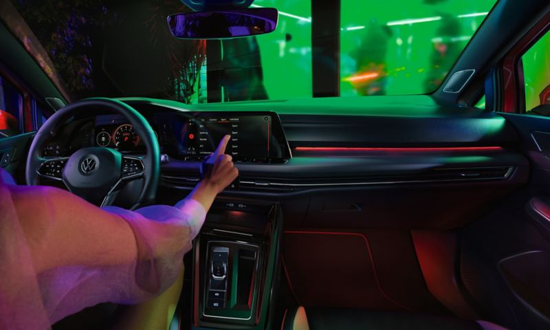"Cockpit of the VW Golf GTI with red background lighting above the glove box and in the footwell, a woman using a touchscreen. "