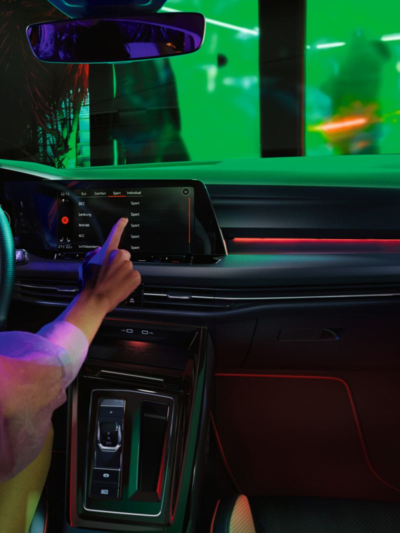 Cockpit des VW Golf GTI mit rotem Ambientelicht über Handschuhfach und im Fußraum, eine Frau bedient den Touchscreen.