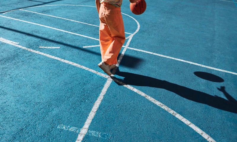 Mann mit orangener Hose spielt auf einem blau gepflasterten Sportplatz Basketball.