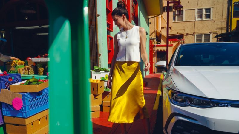 Woman strolls past a fruit stall on a market, a white VW Golf GTE is parked at the roadside.