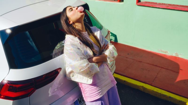 Woman leans on the luggage compartment of a white VW Golf GTE blowing bubble gum.