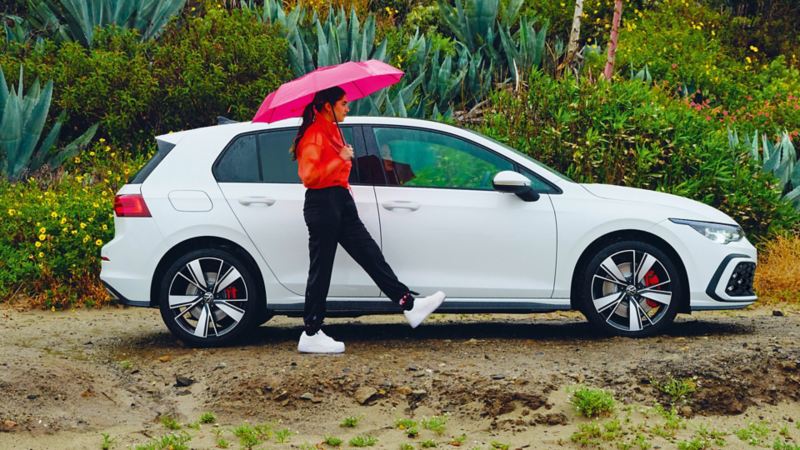 VW Golf GTE in white, side view, stands when it rains in nature, in front of it is a woman with an umbrella