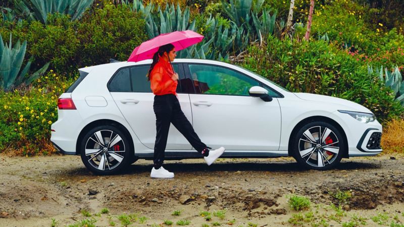 VW Golf GTE in weiss, Seitenansicht, steht, wenn es in der Natur regnet, davor eine Frau mit einem Regenschirm