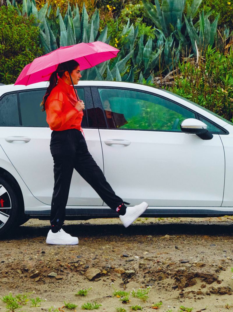 VW Golf GTE in white, side view, stands when it rains in nature, in front of it is a woman with an umbrella