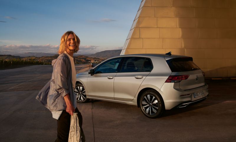 VW Golf MOVE in silber parkt vor einem futuristischen Gebäude, im Vordergrund eine lachende junge Frau