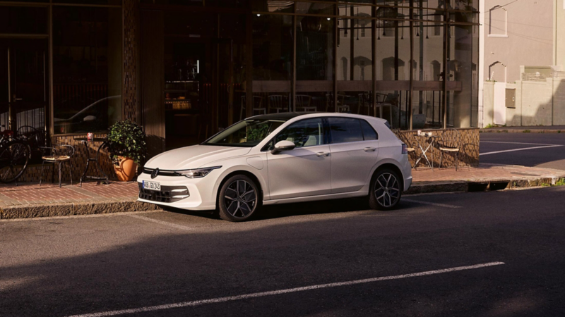 Une VW Golf eHybrid blanche en charge, de profil, garée le long d'un café. Deux hommes se tiennent près de la voiture.