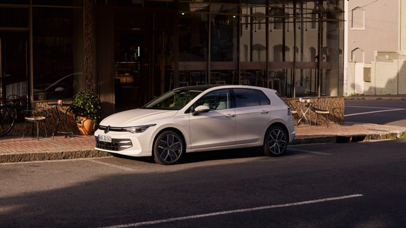 Une VW Golf eHybrid blanche en charge, de profil, garée le long d'un café. Deux hommes se tiennent près de la voiture.