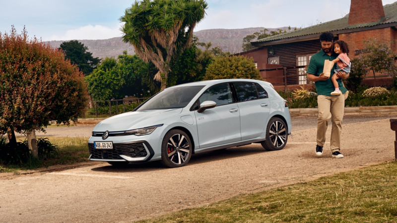 A white VW Golf GTE parked on the road on a drive. Next to the car is a man holding his daughter in his arms.