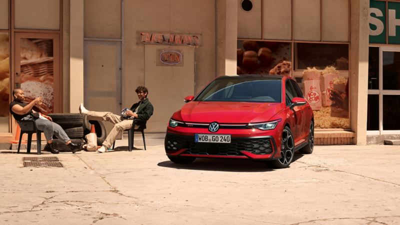 Una Volkswagen Golf GTI rossa ferma davanti a un edificio. Due persone sono sedute vicino all’auto.