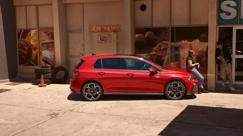 Vue latérale d'une VW Golf GTI rouge devant un magasin. Une femme est assise sur le capot.