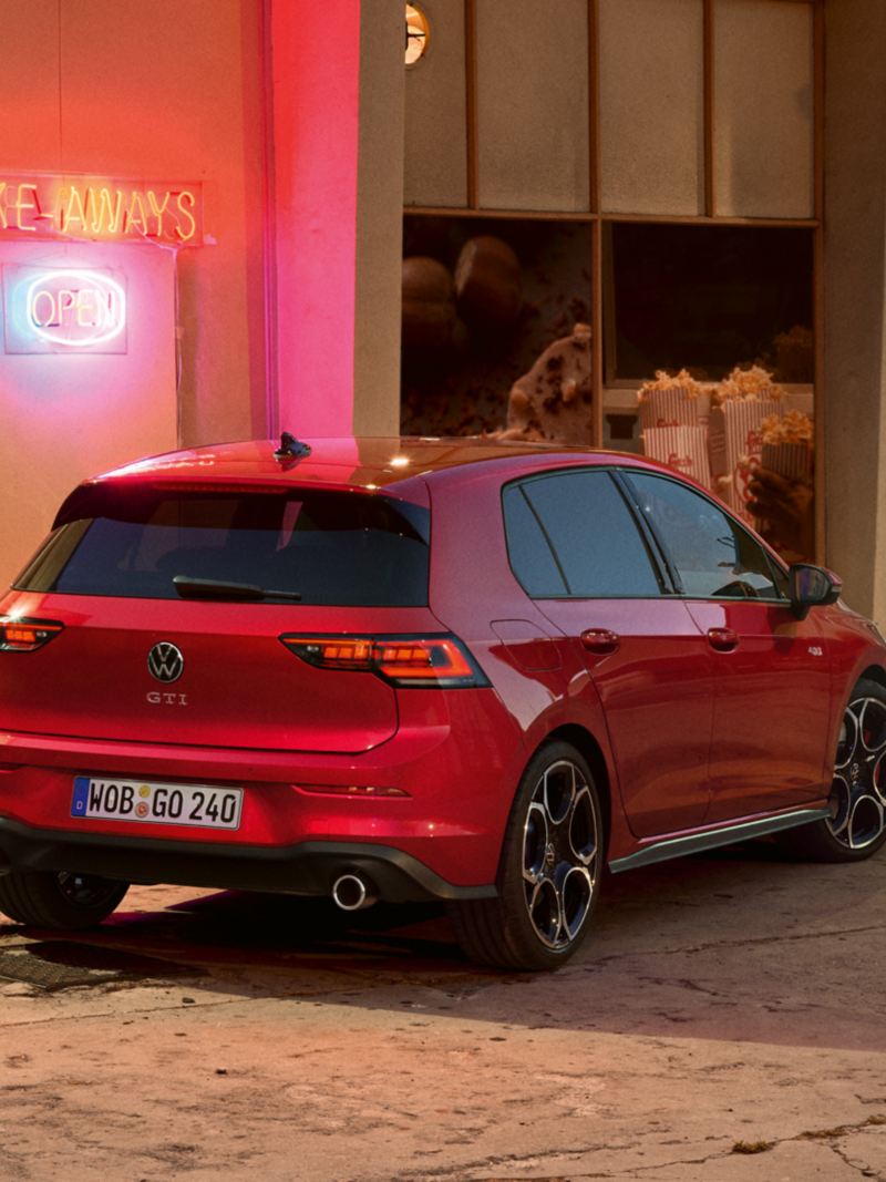 Rear view of a red VW Golf GTI in front of a shop. A man and a woman with snacks in hand approach you.