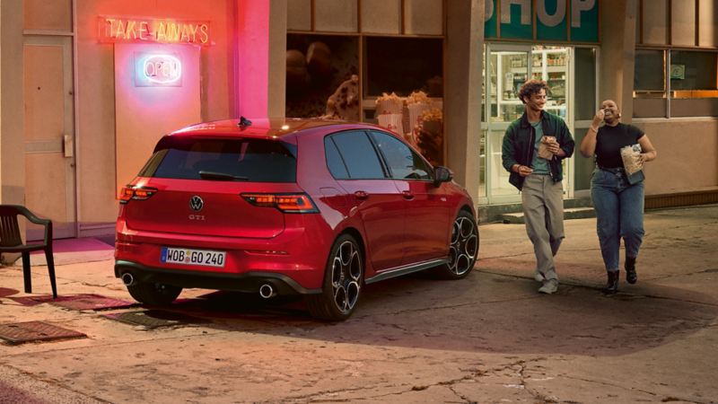 Rear view of a red VW Golf GTI in front of a shop. A man and a woman with snacks in hand approach you.