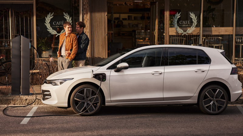 Une VW Golf eHybrid blanche en charge, de profil, garée le long d'un café. Deux hommes se tiennent près de la voiture.