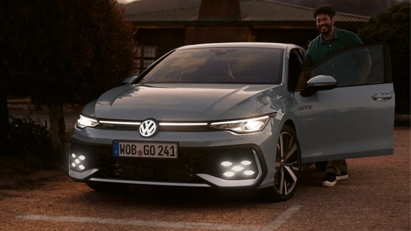 Golf GTE stationnée avec les projecteurs allumés et le logo Volkswagen illuminé. La porte conducteur est ouverte et une personne se tient debout dans l'entrebaillement.