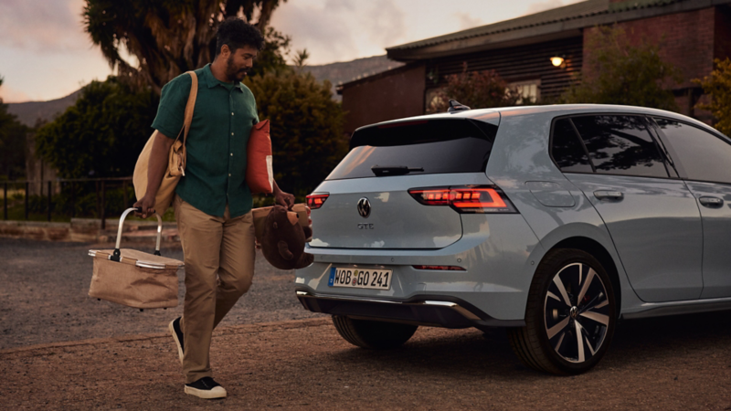 A man in a green t-shirt wants to load some items into the boot of a white VW Golf GTE with its tail light clusters switched on.