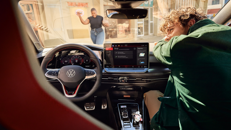 Interior of the Golf with a view from the passenger seat through the windscreen onto the road. A young man stands at the open driver's door.