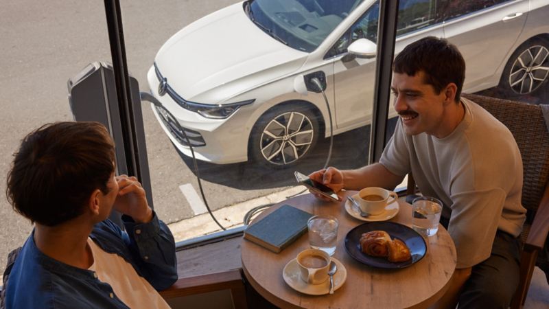 Deux hommes sont assis dans un restaurant, un smartphone à la main. Vue de l'intérieur du restaurant sur la VW Golf garée à l'extérieur, avec le câble de recharge branché.