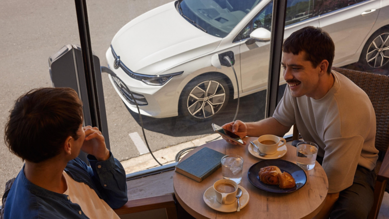 Dos hombres en la mesa de una cafetería. Uno de ellos sostiene su teléfono móvil en la mano. Al fondo se ve un Golf eHybrid cargándose en una estación de carga pública.