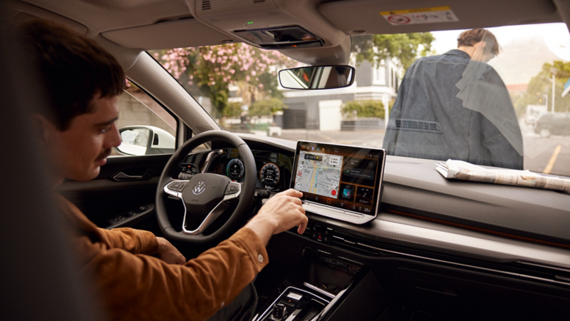 Eine Frau sitzt bei geöffneter Tür auf dem Fahrersitz des gepakrten VW Golf und blickt auf ihr Smartphone.