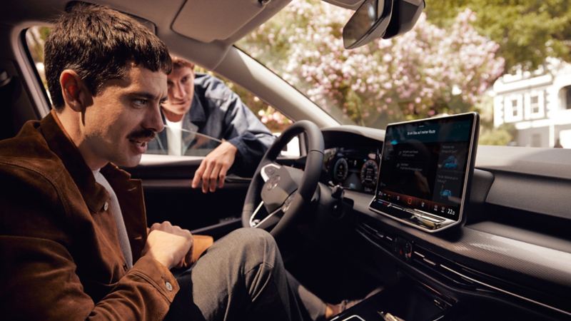 "Innenansicht des VW Golf mit Blick auf den Fahrer, das Cockpit und dem großen Bildschirm. Ein Mann sitz am Lenkrad und ein weiterer Mann ist neben dem Auto. "