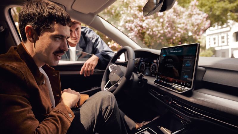 Vue intérieure de la VW Golf mettant l’accent sur le grand écran. Un homme est assis sur le siège du passager avant et regarde en souriant vers le siège du conducteur.