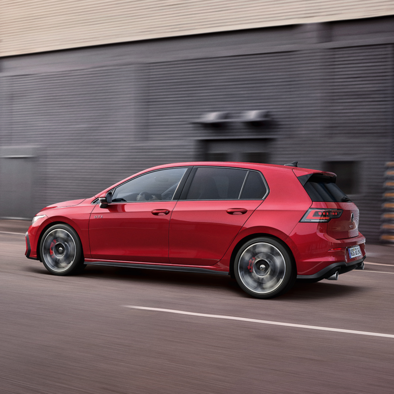 En rød VW Golf GTI set fra siden kører foran en grå bygning.