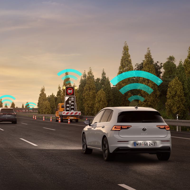 "En hvid VW Golf på en trafikeret motorvej i retning af et vejarbejde. Blå-grønne Wi-Fi-signaler om biler og forhindringer. "