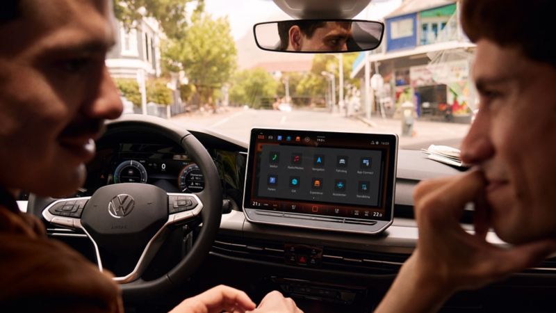 Interior view of the VW Golf with view of the driver, the cockpit and the large display. A man is sitting at the steering wheel and another man is in front of the car.