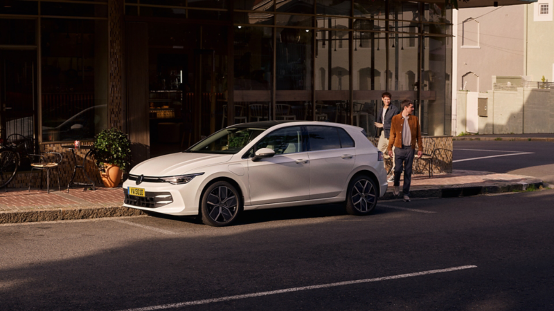 Volkswagen Golf blanche, stationnée sur une rue de ville, avec deux personnes marchant en arrière-plan. 