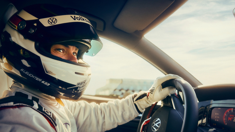 Helmeted Volkswagen GTI race driver grips the steering wheel while driving on track, with digital displays visible inside the cockpit.