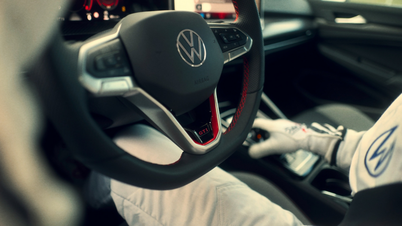 Close-up of Volkswagen GTI steering wheel with red accents as a gloved driver operates the gear selector.
