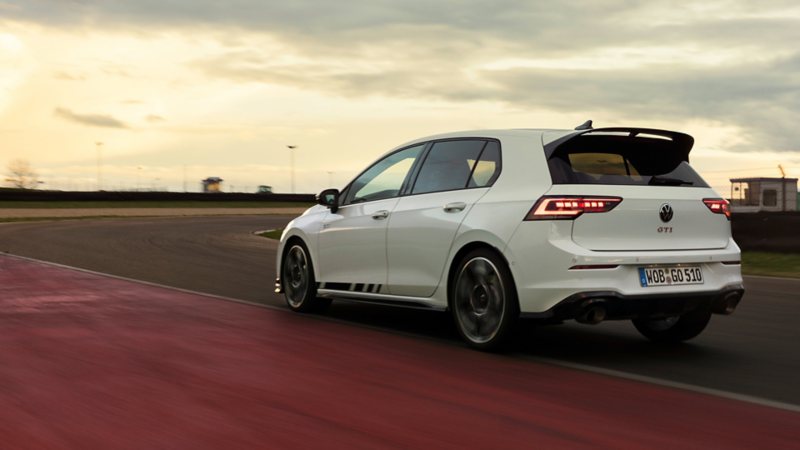 White Volkswagen Golf GTI drives on a racetrack, captured from the rear against a dramatic sunset sky.