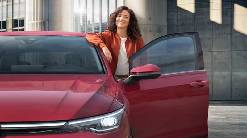 Mujer apoyada en la puerta abierta de un VW Golf rojo está sonriendo.