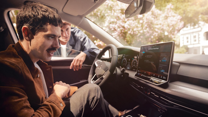 Innenansicht des VW Golf mit Blick auf den Fahrer, das Cockpit und dem großen Bildschirm. Ein Mann sitz am Lenkrad und ein weiterer Mann ist neben dem Auto. 